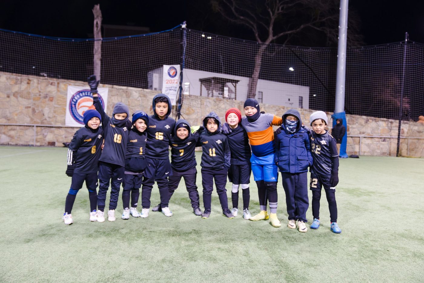 A group of young players in dark jerseys and winter hats smile together on the field at night