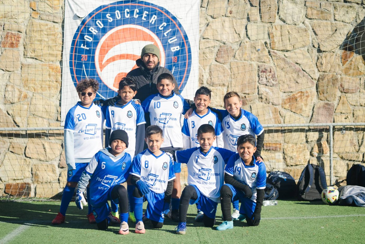 A youth team in blue and white jerseys poses with their coach in front of the Foro Soccer Club banner
